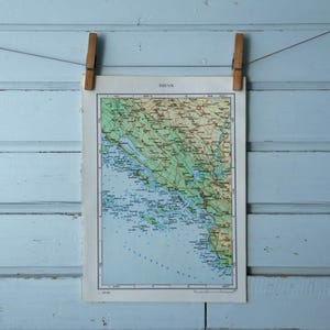 May include: A vintage map of the Brevia region, secured by two wooden pegs against a light blue wooden backdrop. The map displays detailed geographical information with green, brown, and blue hues, highlighting land and water features.