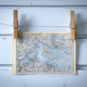 Puede incluir: Una impresión de mapa antiguo, con tonos azules y grises, sujeta por dos pinzas de madera sobre un fondo de madera azul claro. El mapa, suspendido por un alambre fino, muestra un diseño geográfico detallado, creando una exhibición rústica.