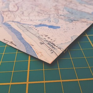 May include: A corner of a vintage map with blue, beige, and brown tones. The map features detailed lines and text, possibly depicting a coastal region. The map is on a green cutting mat.
