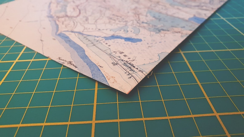 May include: A corner of a vintage map with blue, beige, and brown tones. The map features detailed lines and text, possibly depicting a coastal region. The map is on a green cutting mat.