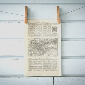 Puede incluir: Un mapa vintage en blanco y negro de una ciudad, impreso en una página de un libro. El mapa está titulado "Viena" y está rodeado de texto en alemán. La página está colgada en una pared de madera blanca con dos pinzas de madera.