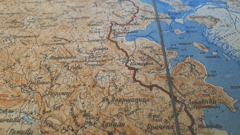 May include: Close-up of a vintage map with detailed topography. The map features shades of brown and blue, with text in a foreign language. A red line marks a route across the landscape. The map appears to be of a mountainous region.