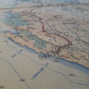 May include: A detailed vintage map with coastal features, including a bay and town labeled "УЛИЦЬ". The map uses a color palette of beige, brown, and blue, with contour lines and text. The map is likely printed on paper.