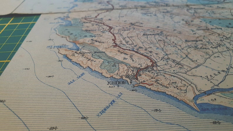 May include: A detailed vintage map with coastal features, including a bay and town labeled "УЛИЦЬ". The map uses a color palette of beige, brown, and blue, with contour lines and text. The map is likely printed on paper.