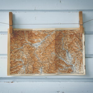 Könnte beinhalten: Eine Vintage-Karte einer Bergregion hängt an einer weißen Holzwand mit Wäscheklammern. Die Karte ist braun und beige mit blauen Linien und Text.
