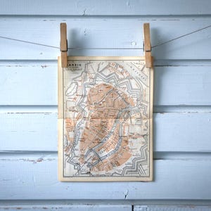 Può includere: Stampa di mappa antica di una città, tenuta da mollette di legno su uno sfondo di legno azzurro. La mappa presenta dettagliati piani della città in tonalità marroni e beige, con testo visibile in alto.