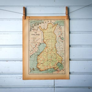 Puede incluir: Un mapa antiguo de Finlandia, exhibido en una pared de madera azul claro. El mapa está impreso en papel envejecido y sujeto por dos pinzas de madera. El mapa presenta información geográfica detallada, con el contorno del país.