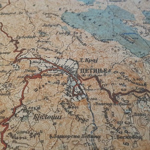 May include: Close-up of a vintage map with detailed topography and place names. The map features a blend of tan, brown, and blue hues, with red lines highlighting roads or borders. The text "ЦЕТИЊЕ" is visible.