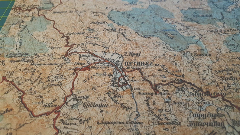 May include: Close-up of a vintage map with detailed topography and place names. The map features a blend of tan, brown, and blue hues, with red lines highlighting roads or borders. The text "ЦЕТИЊЕ" is visible.