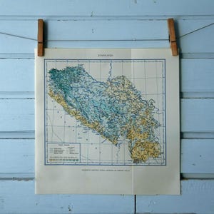 Puede incluir: Impresión de mapa antiguo de una región, probablemente un área geográfica, con tonos azules, verdes y amarillos. El mapa está sujeto con pinzas de madera sobre un fondo de madera azul claro. El mapa tiene líneas de cuadrícula y texto.