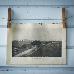 Pode incluir: Uma impressão de paisagem vintage em preto e branco, presa por dois pregadores de madeira em um fio contra um fundo de madeira azul claro. A impressão retrata uma estrada, árvores e edifícios ao longe. O texto "No. 47. Rondo delle" é visível.