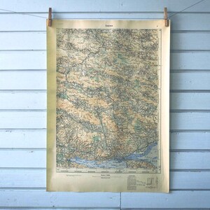 Puede incluir: Impresión de mapa antiguo, colgada en una pared de madera azul claro. El mapa, sujeto con pinzas de madera, muestra detalles geográficos intrincados. El papel envejecido presenta un diseño detallado, con varios colores.