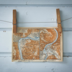 Puede incluir: Un mapa vintage de una ciudad con un río que la atraviesa, colgado en una pared de madera blanca con dos pinzas de madera.