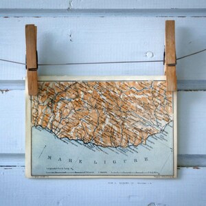 May include: A vintage map print of the Mare Ligure region, held by two wooden clothes pegs against a light blue wooden background. The map features brown and beige tones with blue accents, and the text "MARE LIGURE" is visible at the bottom.