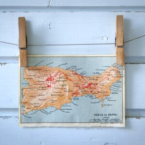 Puede incluir: Un mapa antiguo de Isola di Capri, Italia, sujeto por dos pinzas de madera sobre un fondo de madera azul claro. El mapa presenta tonos rojos y marrones, con la forma de la isla y los nombres de los lugares visibles.