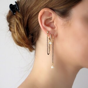 Peut inclure: Une boucle d'oreille en chaîne dorée avec des perles noires et des accents de perles. La boucle d'oreille est portée au lobe de l'oreille et comporte une boucle d'oreille et un clou d'oreille au-dessus.