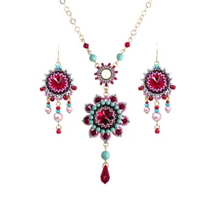 May include: A set of beaded jewelry featuring a necklace and earrings. The necklace has a large, red crystal centerpiece surrounded by smaller pink and turquoise beads. The earrings are similar in design, with smaller red crystals and matching beads.