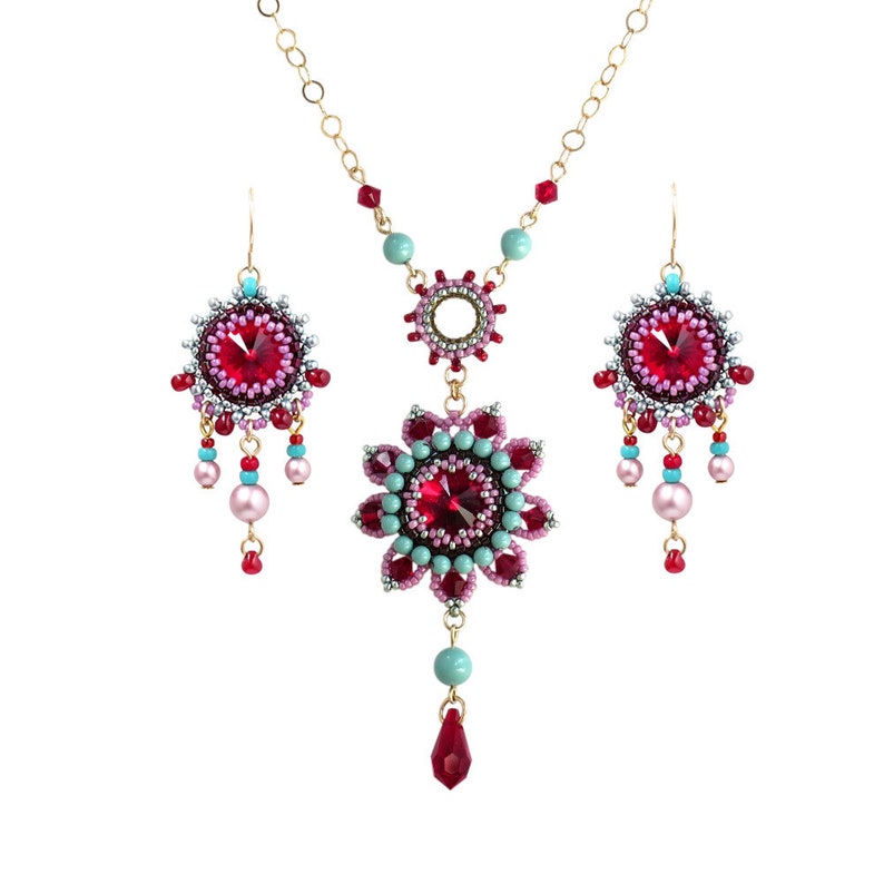 May include: A set of beaded jewelry featuring a necklace and earrings. The necklace has a large, red crystal centerpiece surrounded by smaller pink and turquoise beads. The earrings are similar in design, with smaller red crystals and matching beads.