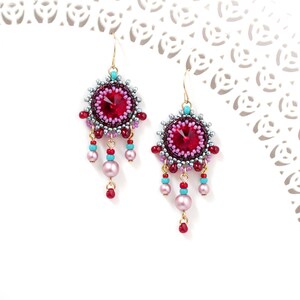 May include: A pair of dangle earrings with a red crystal center surrounded by silver and pink beads. The earrings have a gold hook and dangle with pink and turquoise beads.