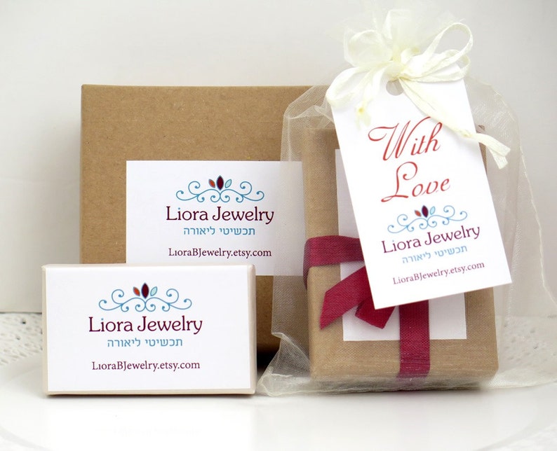 May include: Two brown cardboard boxes with a white label that says "Liora Jewelry" in English and Hebrew. The smaller box is in a clear organza bag with a red ribbon and a white tag that says "With Love" and "Liora Jewelry" in English and Hebrew.