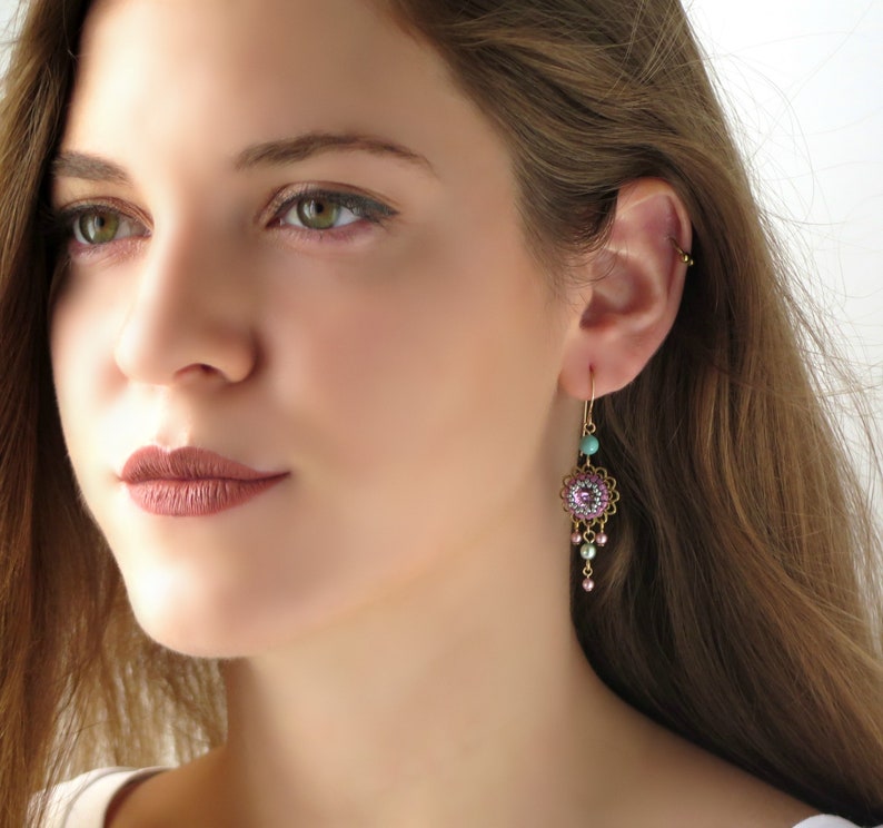 May include: Gold dangle earrings with a pink and turquoise floral design. The earrings feature a round, beaded centerpiece with a pink stone in the center. The earrings are accented with small, pink and turquoise beads.