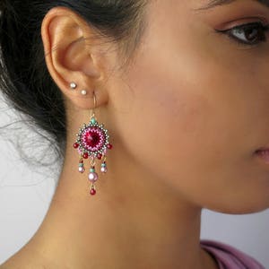 May include: A pair of dangle earrings with a pink and red beaded design. The earrings feature a round, red crystal in the center and a dangling chain of pink and red beads.