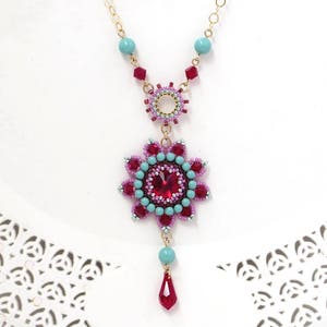 May include: A gold chain necklace with a pink and turquoise beaded pendant featuring a large red crystal. The pendant is a flower shape with a smaller circle in the center.