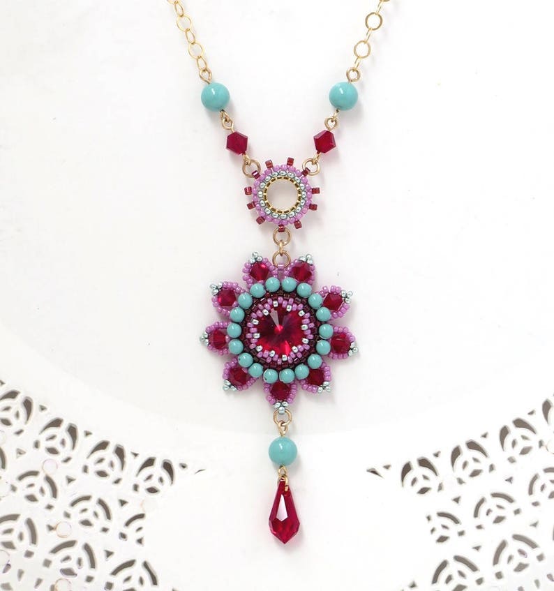 May include: A gold chain necklace with a pink and turquoise beaded pendant featuring a large red crystal. The pendant is a flower shape with a smaller circle in the center.