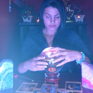May include: A clear glass crystal ball sits on a dark wooden stand, illuminated from within by a warm light. Surrounding the base are multiple rectangular tarot cards arranged in a spread on a table, with a blue-toned light casting a glow.