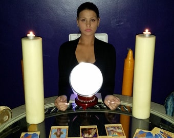 May include: A woman sits at a table with a crystal ball, candles, and tarot cards. The table is covered in a glass top and has a dark wood base. The woman is wearing a black shirt and has dark hair. The background is a dark blue wall with religious artwork.