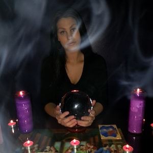 Puede incluir: Una persona sosteniendo una bola de cristal negra, rodeada de velas encendidas y cartas del tarot. El fondo es oscuro con humo, creando una atmósfera mística. Se ven velas moradas y cartas del tarot.