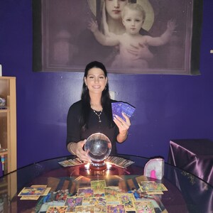 Amanda looking at the camera with a welcoming smile, holding up a selection of tarot cards against a cozy backdrop of glowing candles and crystals.