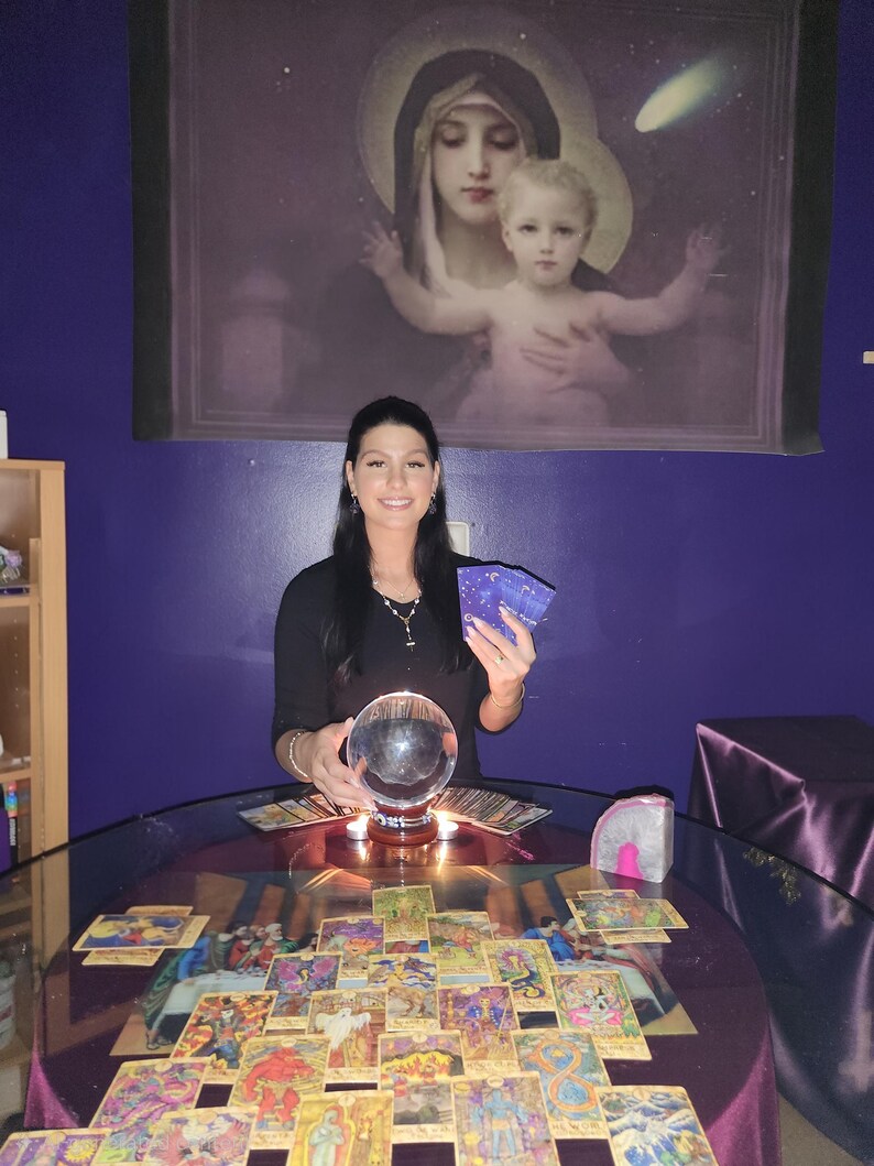 Amanda looking at the camera with a welcoming smile, holding up a selection of tarot cards against a cozy backdrop of glowing candles and crystals.
