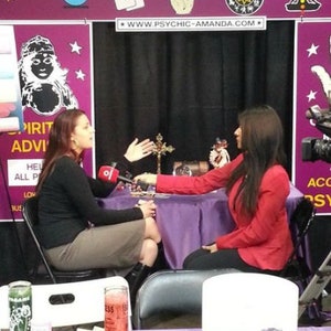 May include: Two people sit at a table with a purple tablecloth. The person on the left is wearing a black shirt and a brown skirt. The person on the right is wearing a red shirt and black pants. The table has a crystal ball, candles, and other items on it. The text "Psychic & Spiritual Amanda" is visible in the background.