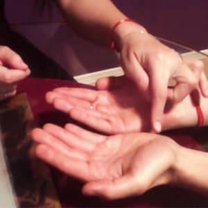Pode incluir: Duas palmas humanas abertas posicionadas lado a lado, com uma mão usando um anel de prata fino no dedo anelar. Um fio vermelho fino está amarrado ao redor do pulso de cada braço e um dedo aponta para o centro de uma das palmas.