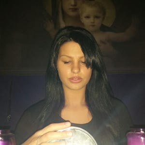 Pode incluir: Uma esfera de vidro transparente segurada por duas mãos, posicionada entre duas velas cilíndricas roxas. A esfera reflete uma luz branca brilhante e uma pintura escura de uma figura segurando uma criança é visível ao fundo, atrás da pessoa.