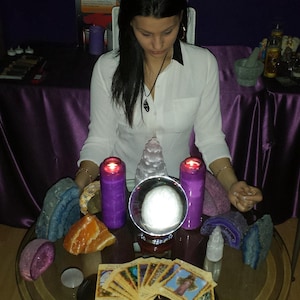 Può includere: Un tavolo in vetro sostiene una sfera di cristallo, due candele a colonna viola, varie rocce geodi colorate e un ventaglio di carte oracolo illustrate. Un poster intitolato CHAKRAS & COLORS è visibile sullo sfondo dietro gli oggetti.