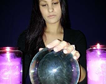 May include: A clear glass crystal ball sits on a dark wooden stand, flanked by two tall cylindrical candles filled with purple wax. Each candle features a raised cross design on the glass surface. Two small tea light candles glow at the base.