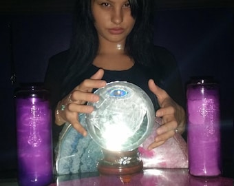 Puede incluir: Una bola de cristal transparente sobre un soporte de madera oscura, situada entre dos velas de pilar moradas altas con diseños de cruces. Una colección de cartas de oráculo rectangulares con ilustraciones coloridas está extendida sobre la mesa.