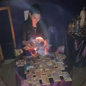 High-angle photo of Amanda analyzing a complex tarot card spread on a table decorated with raw amethyst crystals and a burning stick of incense.
