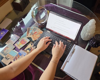 May include: A black laptop with a screen displaying text, surrounded by a clear crystal ball, a deck of tarot cards with various illustrations, a white crystal cluster, and an open spiral notebook with a silver pen on a glass table.