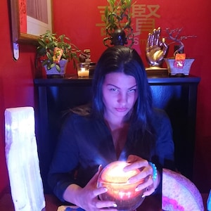 May include: A person with long dark hair sits at a wooden table with a glowing orange crystal ball, surrounded by a spread of rectangular oracle cards. To the left stands a tall white selenite tower, and to the right is a pink geode slice.