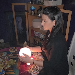 Può includere: Una donna in abito nero è seduta a un tavolo con carte dei tarocchi, tenendo in mano una sfera di cristallo luminosa. Un armadio con oggetti decorativi e uno specchio sono sullo sfondo. La scena è scarsamente illuminata, creando un'atmosfera mistica.