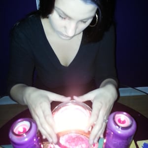 May include: A clear glass sphere glowing with bright light, positioned between two tall purple cylindrical candles with lit pink wicks. The scene includes several small, round, lit pink tea light candles arranged around the central glass sphere.