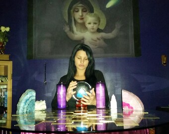 May include: A person with long dark hair sits at a round glass table holding a clear crystal ball. The table features two purple pillar candles, a blue agate slice, a pink agate slice, and a plasma ball lamp. A religious print hangs on the wall.