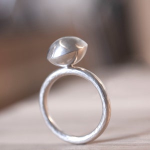Könnte beinhalten: Ein silberner Ring mit einer abgerundeten, kuppelförmigen Oberseite. Der Ring hat eine strukturierte, gehämmerte Oberfläche.
