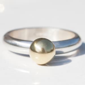 Puede incluir: Un anillo de plata con una bola de oro en la parte superior.