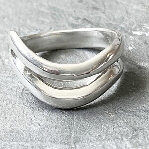 May include: A close-up of a silver ring with a unique, layered design. The ring features two curved bands that flow together, creating a modern and elegant look. The polished metal reflects light, highlighting the ring's smooth, rounded edges. The ring is set against a gray background.