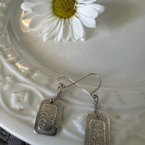 May include: A pair of silver rectangular dangle earrings with a textured, botanical design. The earrings hang from a wire loop and are displayed on a white plate with a daisy flower.