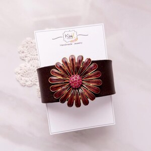 Puede incluir: Una pulsera de cuero marr&oacute;n oscuro con un adorno floral met&aacute;lico grande, p&eacute;talos de color rojo intenso y un centro de brillantes de color rosa intenso. La pulsera se presenta sobre una tarjeta blanca con la inscripci&oacute;n Klay Handmade Jewelry.